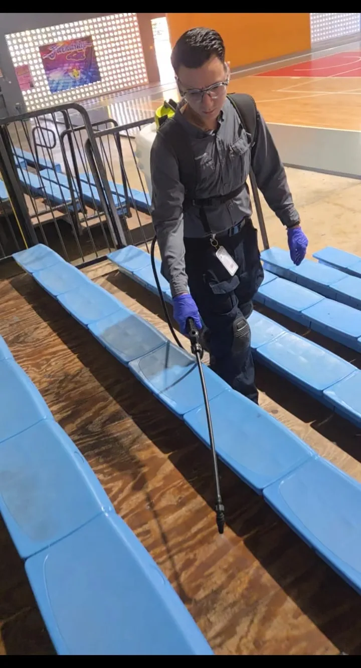 Técnico de Exterminadores PR aplicando tratamiento bajo las gradas de una cancha, con gafas de seguridad, guantes y equipo de aspersión